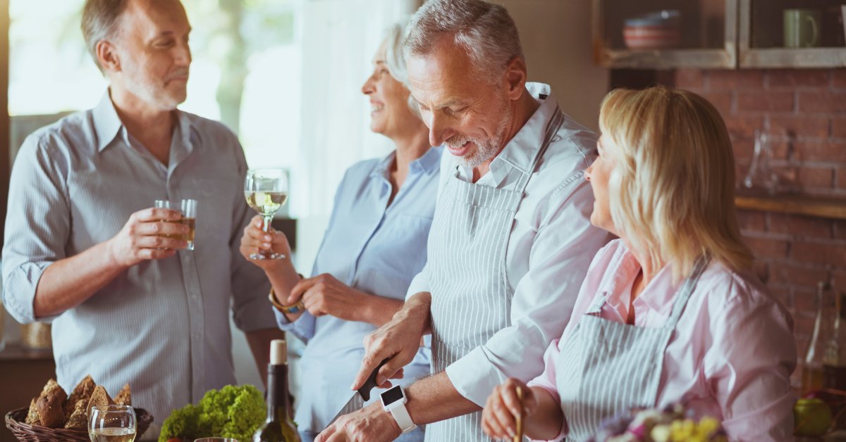Vier ältere Menschen stehen gemeinsam in der Küche, kochen und unterhalten sich entspannt bei einem geselligen Treffen. 