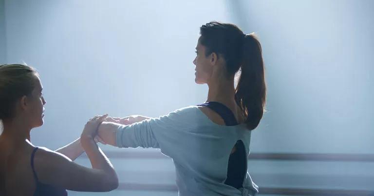 Zwei Frauen, vermutlich Tänzerinnen, üben gemeinsam in einem Tanzstudio mit hellem Licht. Eine Frau führt eine Armbewegung aus, während die andere sie unterstützt. Kein Text vorhanden.