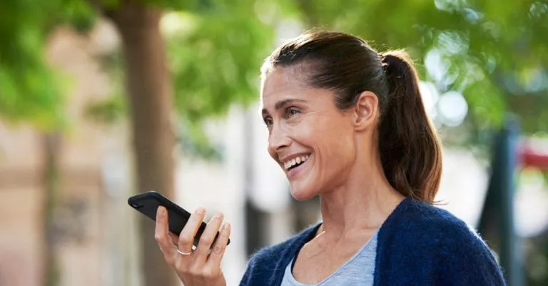 Eine Frau mit Pferdeschwanz hält ein Smartphone in der Hand, scheinbar sprechend oder hörend, während sie in einem sonnigen, grünen Aussenbereich steht. Kein Hörgerät oder Text sichtbar.