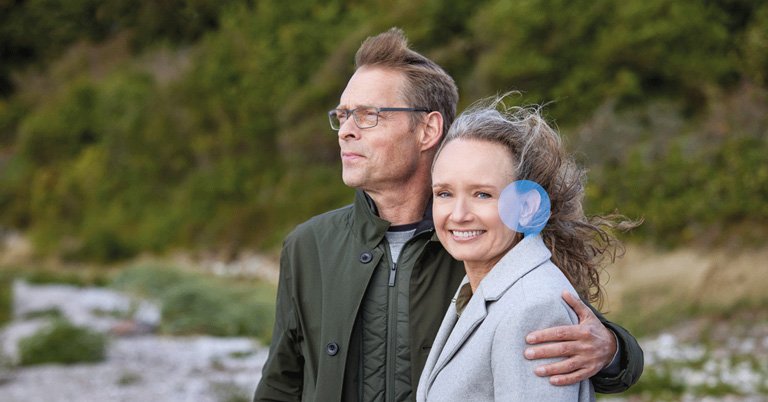 Ein Mann und eine Frau stehen eng umarmt in einer natürlichen Umgebung mit Bäumen und Kiesboden. Die Frau trägt ein diskretes blaues Hörgerät hinter ihrem Ohr.