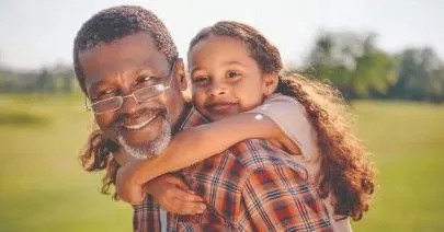Älterer Mann trägt lachendes Enkelkind auf dem Rücken in einer sonnigen Parklandschaft
