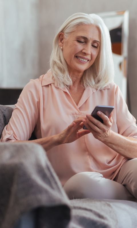 Lächelnde ältere Frau mit schulterlangem grauem Haar sitzt auf einem Sofa und blickt auf ihr Smartphone, im Hintergrund steht eine grüne Zimmerpflanze.