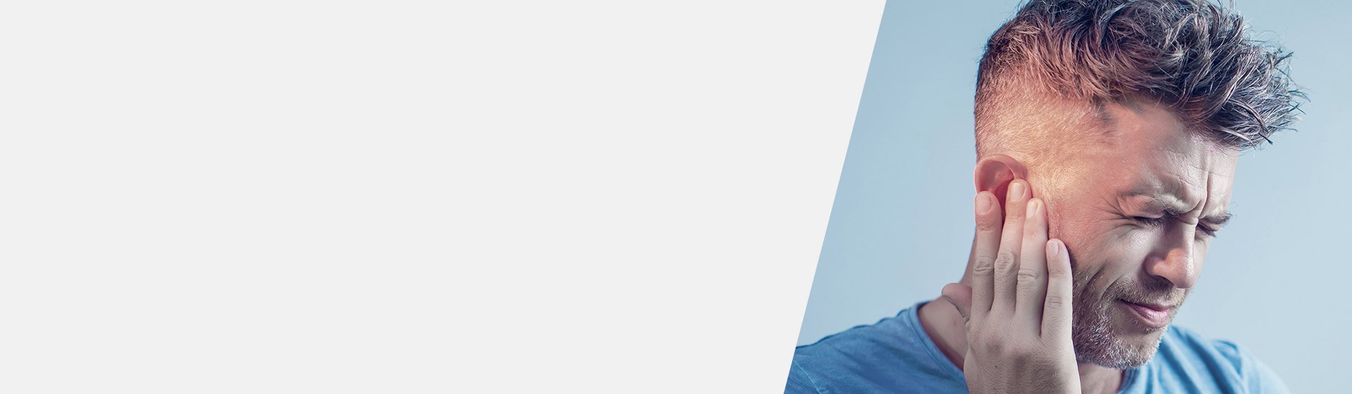 Ein Mann mit kurzen Haaren berührt sein Ohr mit der Hand. Der Hintergrund ist blau und schlicht. Das Bild könnte auf Hörprobleme oder eine Hörtest-Situation in einer Hörklinik hinweisen.