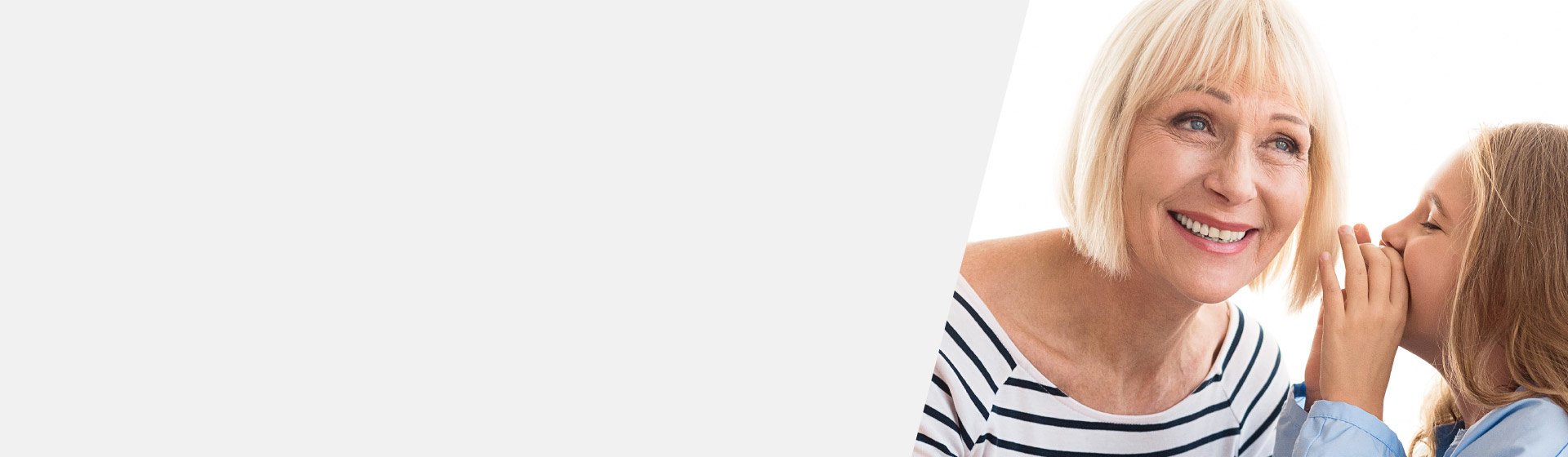Eine ältere Frau in gestreiftem Shirt lehnt sich zu einem Kind, das ihr ins Ohr flüstert; heller Hintergrund suggeriert eine freundliche, familiäre Atmosphäre. Kein Text oder erkennbares Hörgerät sichtbar.