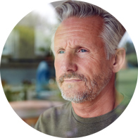 Ein grauhaariger Mann trägt ein grünes Oberteil, sitzt in einem hellen Raum und ist vor einem unscharfen Hintergrund positioniert. Der Fokus liegt auf Hörgesundheit; keine erkennbaren Hörgeräte.