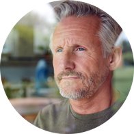 Ein grauhaariger Mann trägt ein grünes Oberteil, sitzt in einem hellen Raum und ist vor einem unscharfen Hintergrund positioniert. Der Fokus liegt auf Hörgesundheit; keine erkennbaren Hörgeräte.