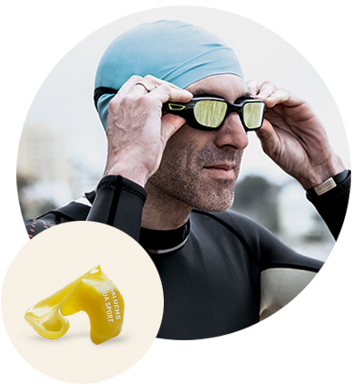 Ein Schwimmer in Neoprenanzug und blauer Badekappe justiert seine Schwimmbrille; im Vordergrund wird ein gelber, individuell angepasster Schwimmschutz für die Ohren gezeigt. Text: keiner.