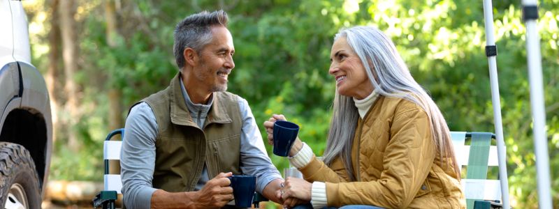 Modent par sidder smilende og drikker kaffe udenfor. I baggrunden ses skovområde og deres camper.