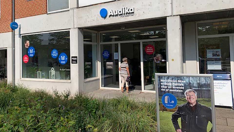 Audika hørerådgivningsklinik med glasindgang og tydelig skiltning, herunder reklame for Oticon More høreapparater: ’Med Oticon More bliver jeg igen en levende del af samtalen.’ En person går ind.