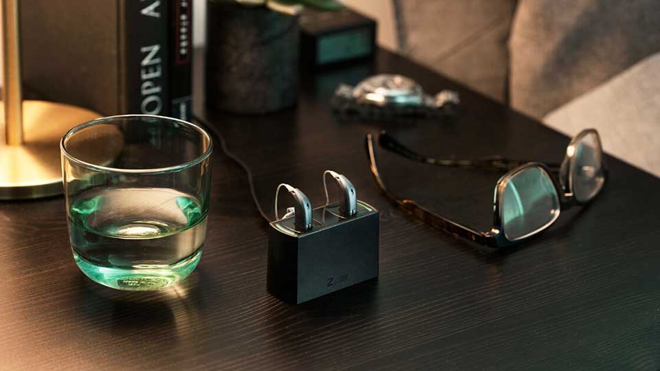 To høreapparater oplader i en sort ladestation på et mørkt træbord, omgivet af et glas vand, briller og bøger. Teksten ’Z...’ kan skimtes på ladestationen.