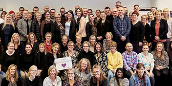 En stor gruppe voksne mennesker smiler og poserer sammen i et lyst lokale. En person holder et skilt med teksten: ’VI ELSKER LYD’ og en lilla hjerteillustration.