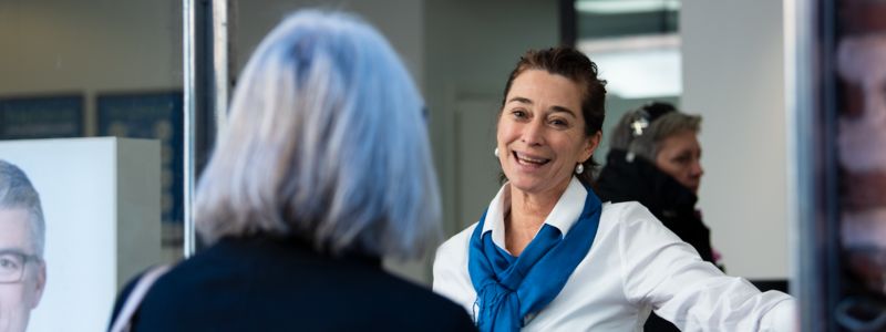 En Audika-ansat, en mørkhåret, smilende kvinde, tager imod en kunde, som vi ser bagfra. 