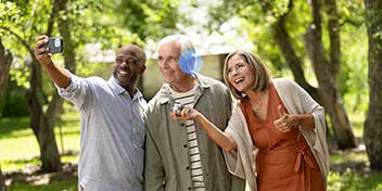 Tre personer tager en selfie i en grøn park. En mand i midten bærer en diskret høreapparat fra Oticon bag øret, mens de øvrige personer smiler og griner.