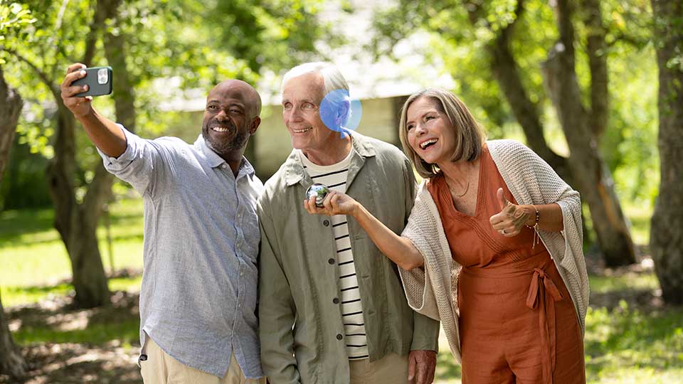 Tre personer står i en have og tager en selfie. En kvinde viser en lille genstand, muligvis et høreapparat, mens omgivelserne består af træer og solskin.