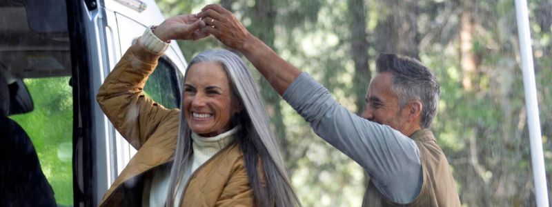 Moden kvinde og mand danser i regnen uden for deres autocamper, mens de griner og smiler.