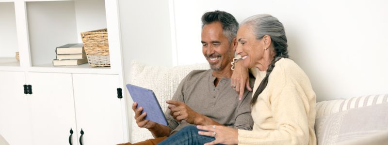 Et modent par sidder i en sofa i en lys stue og kigger sammen på en tablet, som manden holder. De ser ud, som om de ser noget sjovt og har det hyggeligt.