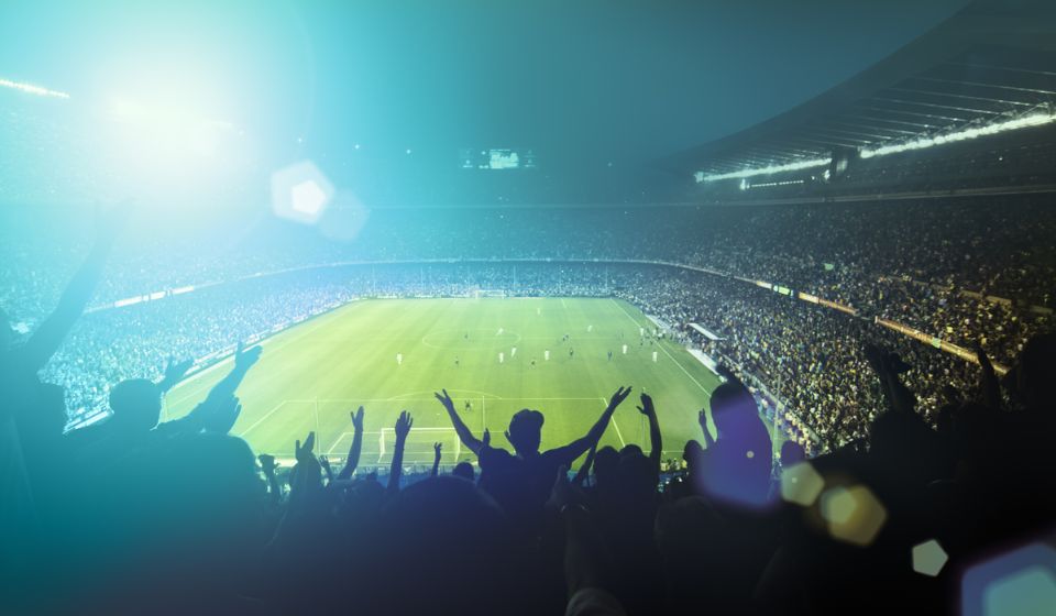 Fodboldstadion med fodboldkamp og heppende fans. Det er mørkt, og der er kunstigt lys på banen.