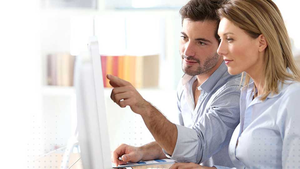 En computer viser information, mens to personer, en mand og en kvinde, interagerer. Manden peger på skærmen i et lyst rum med boghylder i baggrunden.