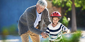 En ældre mand støtter et barn med rød cykelhjelm, mens barnet lærer at cykle på en grøn cykel; udendørs omgivelser med træer og en blå væg i baggrunden.