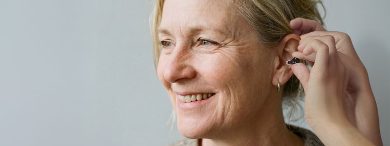 Man ser en smilende kvinde fra siden og et par hænder, der sætter et Oticon Zeal-høreapparat i kvindens øre.