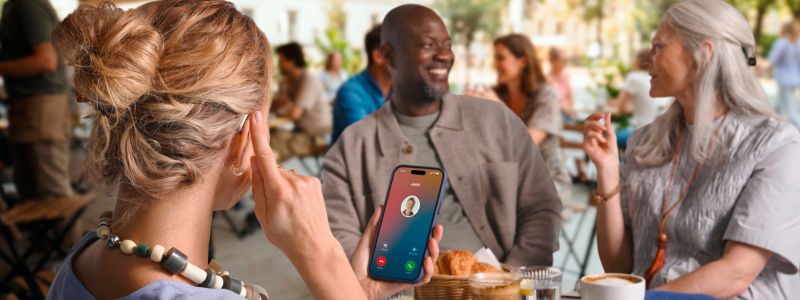 Kvinde sidder på cafe med to venner, en mand og en kvinde, og tager sin telefon ved at dappe på sit øre. Hun har telefonen i venstre hånd og kan nu streame opkaldet.