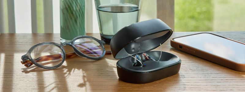 Oticon Zeal-høreapparater ligger i deres æske på et træbord, hvor man også ser et par briller, et glas vand og en smartphone.