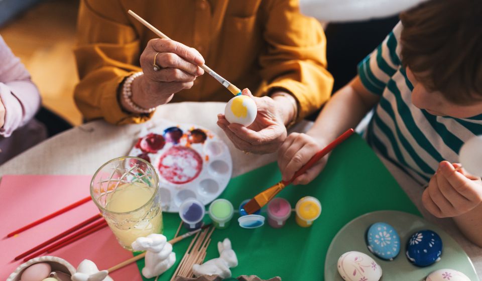 En kvinde sidder og maler et påskeæg sammen med to børn. Bordet er fuld af farver og æg. 