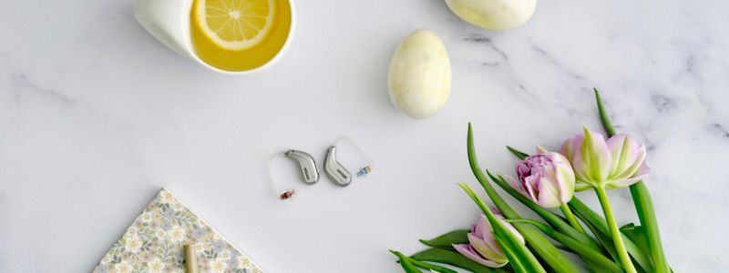 Oticon Intent-høreapparater på et marmorbord, hvor der også er to æg, et glas te med citron og en buket tulipaner. 