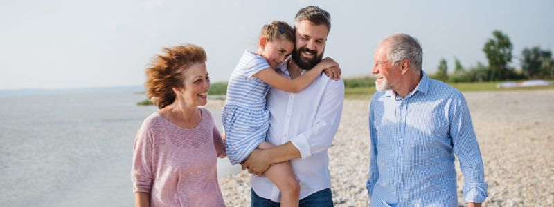 Familie - bedsteforældre, søn og barnebarn - går langs stranden. Solen skinner, og man kan se, at det blæser lidt. 