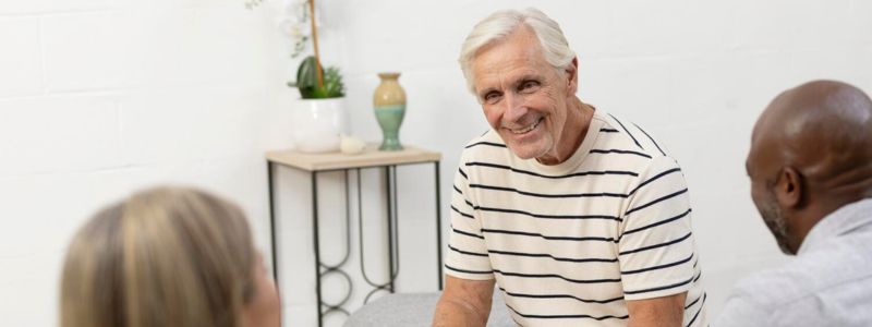 Ældre mand sidder i en sofa og taler med sine to venner, en kvinde og en mand. 