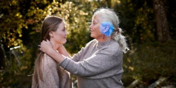 Ældre kvinde står front mod ung pige og holder hende på skuldrene. De er udenfor, og solen skinner. 