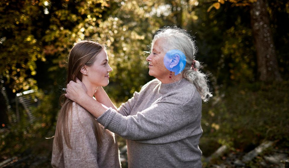 Ældre kvinde står front mod ung pige og holder hende på skuldrene. De er udenfor, og solen skinner. 