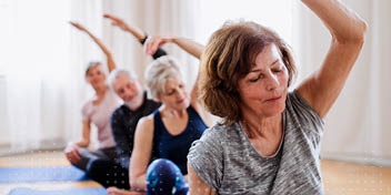 Fire ældre mennesker strækker sig i en yogaklasse i et lyst rum.
