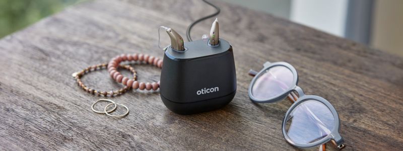 Et sæt Oticon Intent-høreapparater sidder i opladeren, som står på et bord, hvor man også kan se et par briller og nogle armbånd.