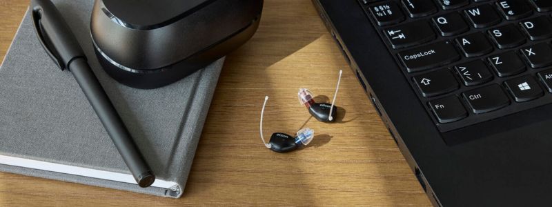 Et par Oticon Zeal-høreapparater ligger på et skrivebord, hvor man også kan se et udsnit af en computer, en notesbog, en kuglepen og etuiet/opladeren til høreapparaterne.