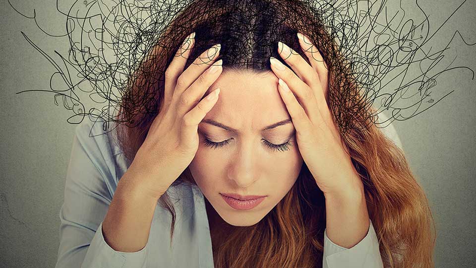 En person med langt, brunt hår holder hovedet med hænderne, omgivet af tegnede, kaotiske streger, hvilket illustrerer stress eller tinnitus. Baggrunden er enkel og grå.