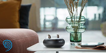 To høreapparater fra Oticon lades i en sort opladningsstation på et hvidt stuebord. Ved siden af står en glasvase med tørrede blomster og en mobiltelefon.