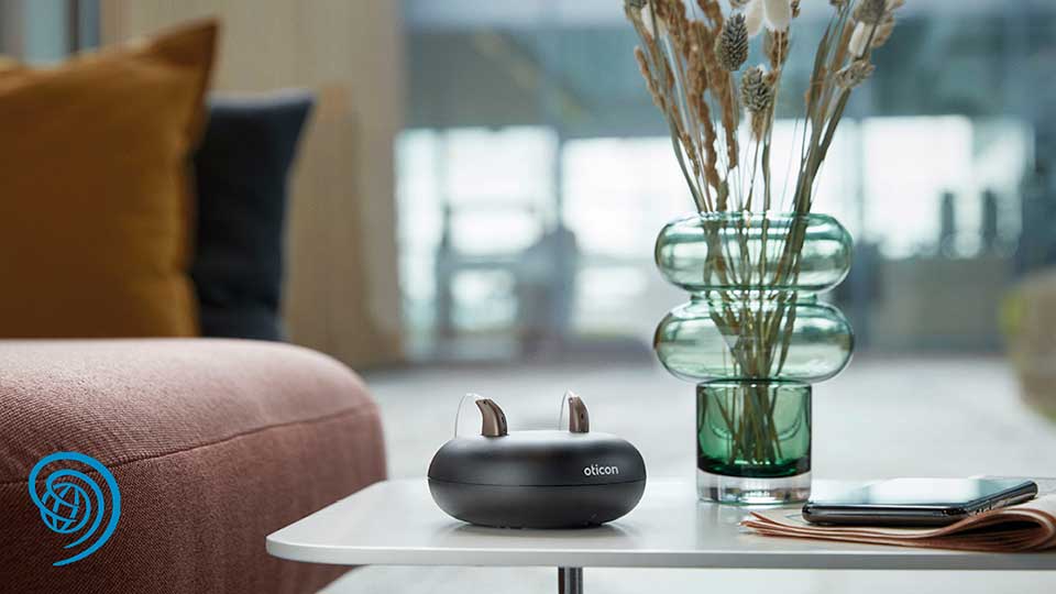 Genopladelige Oticon-høreapparater placeret i en bordoplader ved siden af en grøn glasvase og en smartphone på et stilrent bord i en moderne stue.