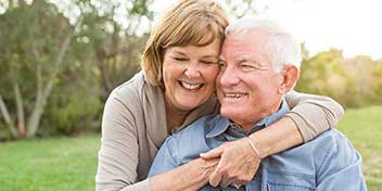 En ældre kvinde omfavner en ældre mand bagfra, mens de er udendørs i en have.