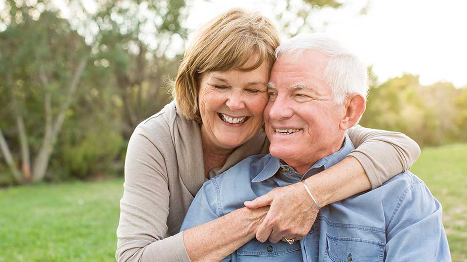 Et ældre par sidder tæt i en park, hvor kvinden omfavner manden bagfra. De er omgivet af grønt græs og træer under naturligt dagslys. Ingen tekst eller høreapparater synlige.