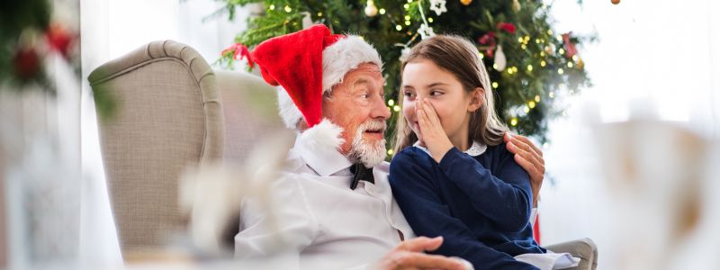 Barnebarn hvisker i øret på sin bedstefar, der sidder i en lænestol med nissehue på. I baggrunden kan man se et pyntet juletræ.