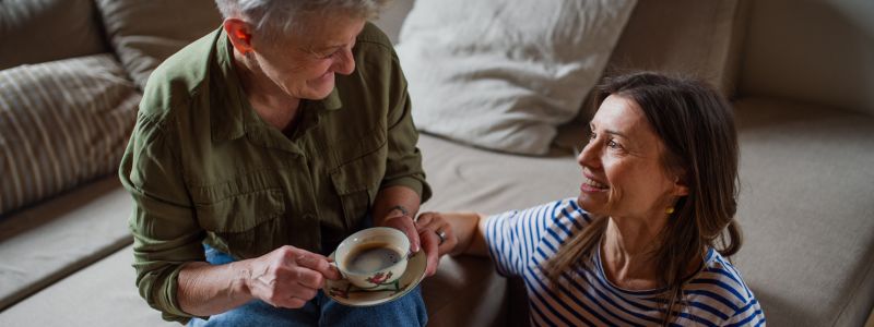 Kvinde sidder i sofaen med en kop te og taler med en yngre kvinde, der sidder på gulvet. De har øjenkontakt og smiler til hinanden.