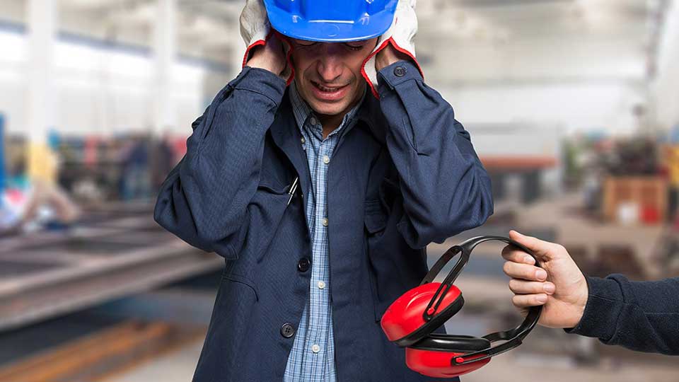En fabrikarbejder med blå hjelm holder hænderne på ørerne, mens en anden person rækker høreværn frem. Baggrunden viser en lys industribygning med slørede maskiner og arbejdsområder.