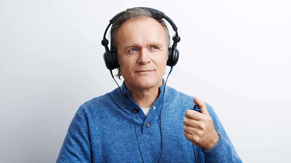 En mand iført hovedtelefoner trykker på en håndholdt knap under en høretest i en hvid klinisk baggrund, muligvis hos en Audika høreklinik.