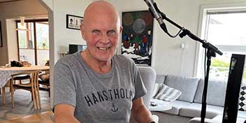 Jens Sørensen sidder ved en mikrofon i en stue med lysindfald og kunst på væggen.