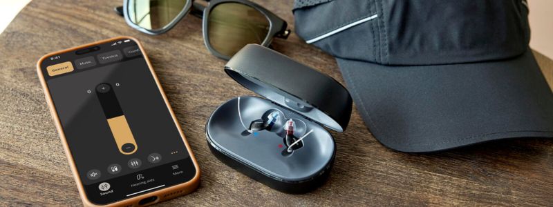 Oticon Zeal-høreapparater ligger i oplader på et bord med smartphone, kasket og solbriller. 