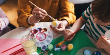 En kvinde sidder og maler et påskeæg sammen med to børn. Bordet er fuld af farver og æg. 