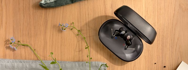 Oticon Zeal-høreapparater i deres oplader/æske på et træbord, hvor man kan se blomster og nogle havehandsker.
