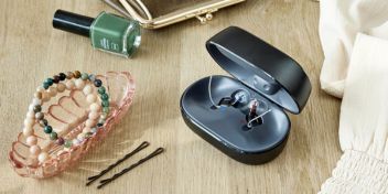 Et sæt Oticon Zeal-høreapparater sidder i opladeren, som står på et bord, hvor man også kan se en grøn neglelak, nogle armbånd og nogle hårnåle.