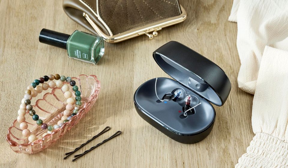 Et sæt Oticon Zeal-høreapparater sidder i opladeren, som står på et bord, hvor man også kan se en grøn neglelak, nogle armbånd og nogle hårnåle.